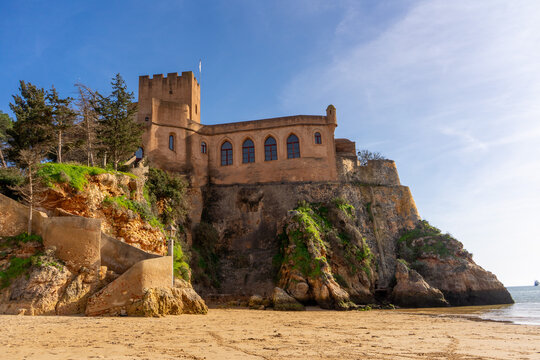 Sandiger K&uuml;stenbereich im Vordergrund mit Blick auf das Castelo de S&atilde;o Jo&atilde;o do Arade, historische Festung auf einem Felsen bei Ferragudo