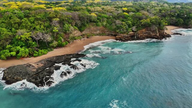Aerial view of the rugged coastline where turquoise waters crash against dark rocks and meet a sandy beach fringed by lush green forest, Tambor, Costa Rica.