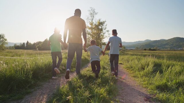 Rear view of an African American father and his three boys walking together holding hands along a meadow path