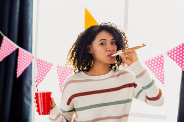 Obraz premium Young mixed race woman at home celebrating party with party horn and cake in a bright living room