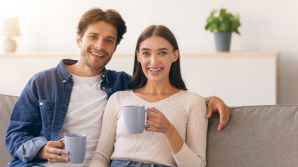 A man and woman sit on a sofa in a living room. They smile and hold mugs in their hands. Sunlight...