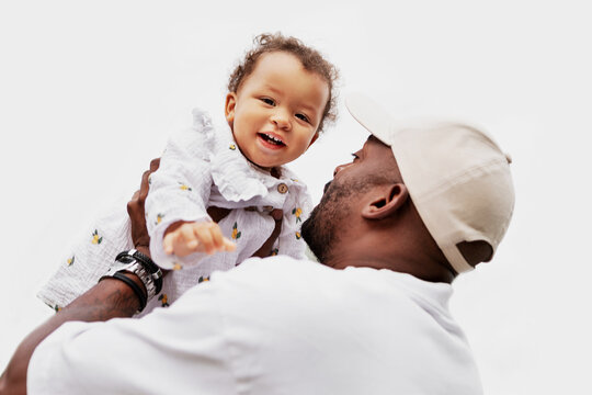 Joyful African American cool dad lifting laughing baby girl up, playful tender moment against white background, happy fatherhood lifestyle portrait