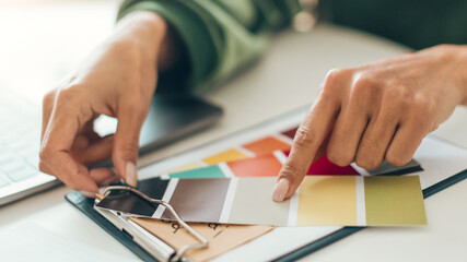 Fototapeta premium A person is selecting paint color samples from a binder on a desk. The workspace includes a laptop and a variety of color options. The individual is focused on making choices for a home project.
