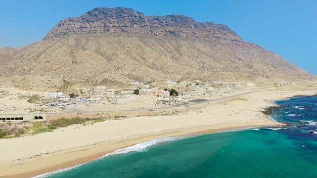Aerial view of buildings and a sandy beach situated between a massive mountain and the turquoise waters of the Gulf of Aden, Car Madow, Somalia.