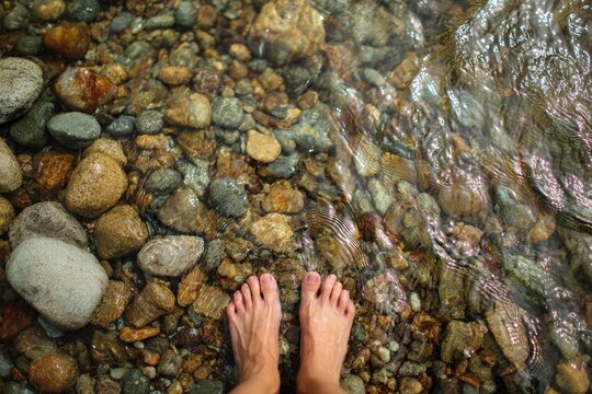 Bare feet submerged in shallow, clear water over colorful, smooth river stones