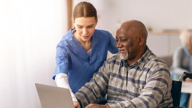A young woman in blue scrubs teaches an older man how to use a laptop in a care facility. They are both engaged in the activity. Other residents are visible in the background.