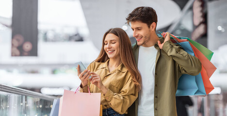 A young couple is at a mall looking at their phone while holding shopping bags. They appear happy and engaged with each other. The mall is busy with other shoppers around them.