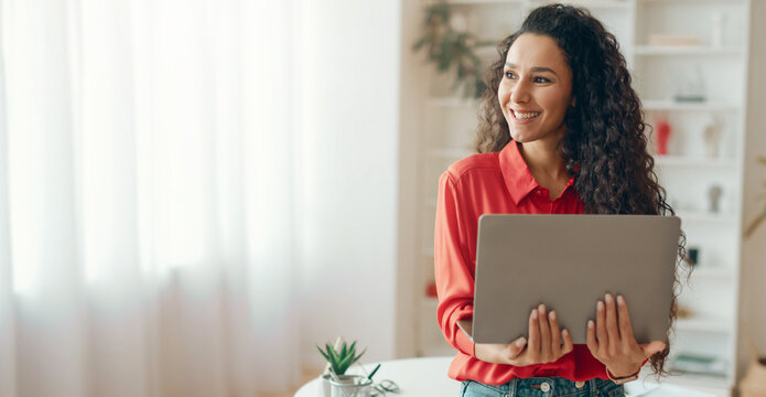 A woman stands in a bright room with plants. She holds a laptop close and smiles. Sunlight comes through the window and lights up the space. She wears a red shirt and looks happy.