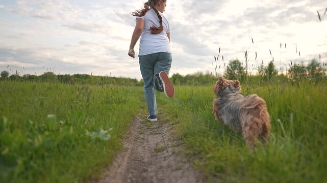 Girl in motion runs with dog through open meadow path. Motion flows in every step as girl leads dog. Outdoor nature wraps them in light. Girl and dog explore wide green space in full summer movement.