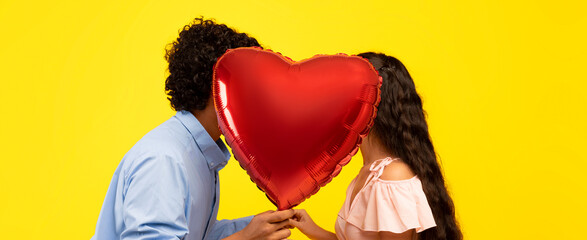 A couple stands close together, holding a heart-shaped balloon that covers their faces. They share a moment of connection. The background is bright yellow, adding to the cheerful scene. © Prostock-studio