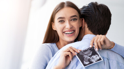 A woman holds an ultrasound image while smiling at the camera. A man stands behind her with his arm around her. They are celebrating their pregnancy together. © Prostock-studio