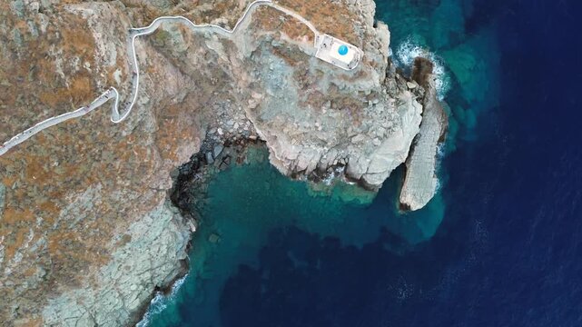 High quality cinematic drone footage looking straight down at the white Church of the Seven Martyrs and the zigzag stone path on a rocky cliff surrounded by turquoise Aegean sea in Sifnos