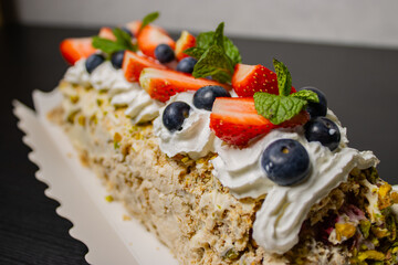 Dessert cake roll topped with whipped cream, strawberry, blueberry, mint, and crushed pistachio on dark table