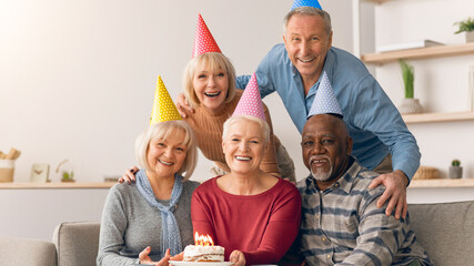 Obraz premium Friends gather together to celebrate a special occasion. They wear colorful party hats and smile while holding a cake with candles. The group enjoys their time in a cozy and welcoming space.