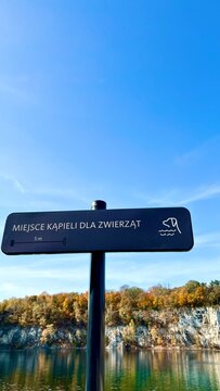 Zalew Zakrzowek, Krakow, Poland. Sign reading Miejsce kapieli dla zwierzat stands before calm blue water, light rocky cliffs, and autumn trees under a clear sunny sky