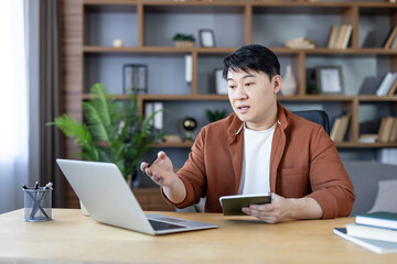 Asian man sitting at desk, holding pen and notebook, talking and gesturing while video conferencing on laptop from home office with bookshelves in background © Liubomir