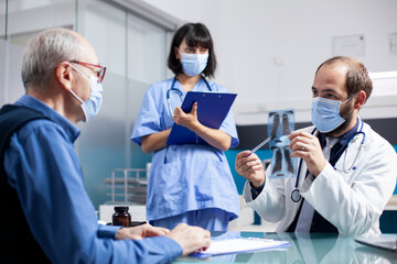 Obraz premium Doctor explaining xray results to elderly man as female medic assistant takes notes during medical appointment. Nurse with clipboard standing next to physician showing radiograph to pensioner patient.