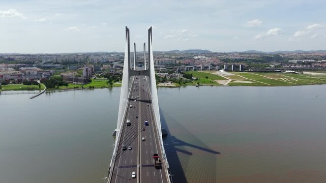Aerial drone footage flying towards the majestic Vasco da Gama Bridge in Lisbon, Portugal. Cars and trucks cross the cablestayed bridge over the Tagus River on a bright, sunny day. Cityscape visible.