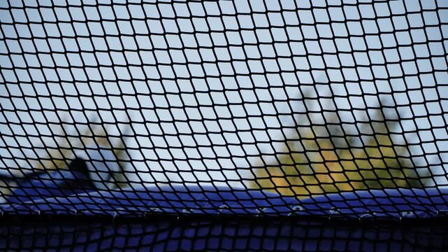 Person jumping on trampoline seen through safety net against blue sky