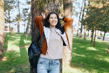 Woman outdoors park tree casual jacket with backpack smiling in sunlit greenery relaxed outdoor...