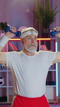 Healthy lifestyle concept where likable confident sportive grey-bearded mature sportsman raising hands with dumbbells when sports training at home in the evening