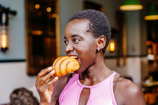 Black woman in pink sleeveless top eating croissant in cafe close up. Female customer looking aside while having fresh pastry, warm bakery interior, breakfast break, urban food lifestyle,
