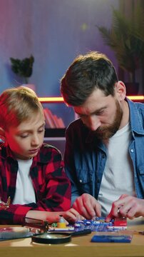 Front view of good-looking smiling happy loving 35-aged bearded dad which sitting at the table together with his 12-aged good-natured son collecting details of constructor
