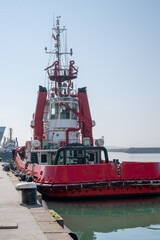 a bright red and white tugboat moored at a concrete pier, showcasing its heavy black rubber fendering, thick mooring lines, and towering bridge structure under a clear, pale blue sky. © Andrey