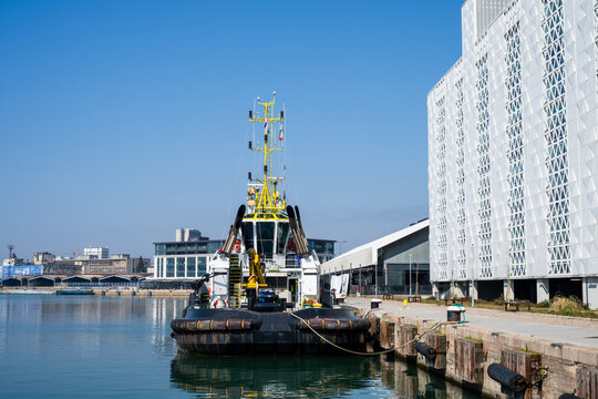 a black, white, and yellow tugboat moored alongside a concrete pier with rubber tire fenders, situated next to a modern white textured building in a calm harbor under a clear blue sky.