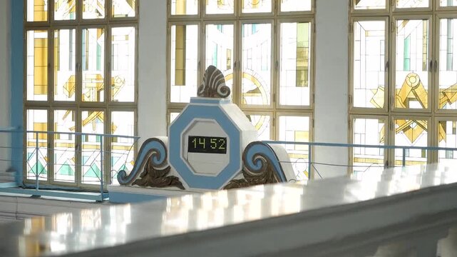 Vintage digital clock displaying time fourteen fifty-two mounted on ornamental pedestal against large industrial windows with geometric stained glass patterns indoors
