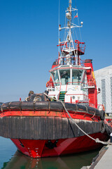 a red and white tugboat moored at a concrete pier, featuring heavy black rubber fenders on its bow and a tall mast equipped with radar and antennas, set against a clear blue sky. © Andrey