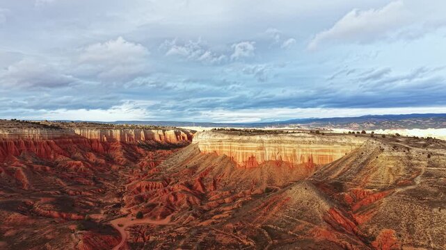 The view shows a wide canyon filled with various layers of rock. The sky is cloudy, creating shadows on the canyon walls. Some green patches are visible in the canyon.