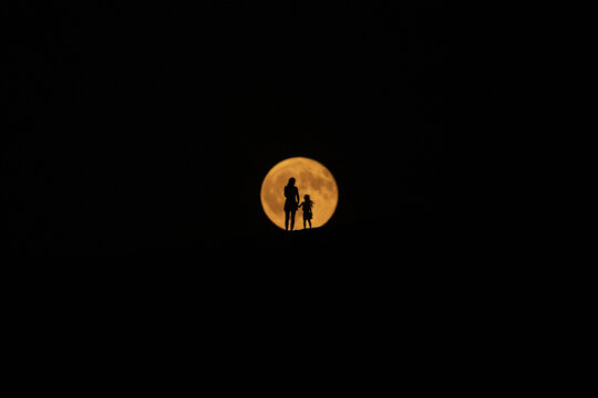 View of silhouetted figures of a parent and child stand against a golden orb of the moon, casting a warm glow in the darkness, Dune du Pilat, Nouvelle-Aquitaine, France.