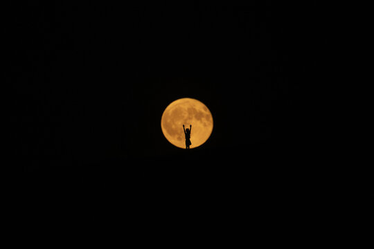 View of a lone figure silhouetted against a colossal, glowing orange moon, arms raised in a gesture of triumph or supplication, Dune du Pilat, Nouvelle-Aquitaine, France.
