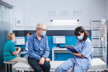 Obraz premium Nurse practitioner taking notes at consultation with senior man in hospital office. Elderly male patient attending healthcare examination with medical assistant during covid 19 pandemic.