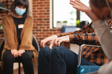 Fototapeta premium Closeup of male person touching his forehead, seated near elderly wheelchair user in brick wall room. Selective focus on hands of man in wheelchair as young woman in face mask sitting nearby.