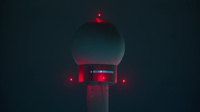 Static close-up shot of an airport radar tower at night. The radome and structure are illuminated by blinking red aviation warning lights against a dark, moody sky