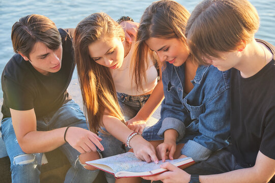Group of young friends planning summer travel adventure, reading paper map for road trip navigation, discussing route with togetherness and teamwork