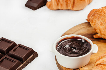 Chocolate spread bowl with croissants and dark chocolate pieces on light background with copy space
