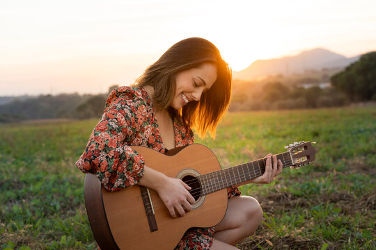 Smiling young woman playing guitar on meadow during sunset