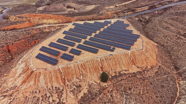 Solar panels cover a hillside in a remote location, arranged in rows under bright sunlight. The surrounding landscape shows dry earth and minimal vegetation.