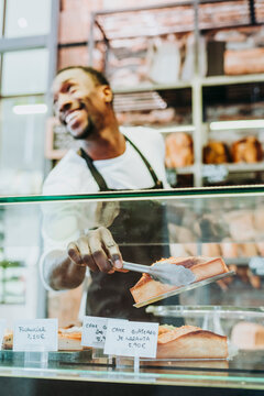 Smiling man working in a bakery taking a piece of cake with a tongs