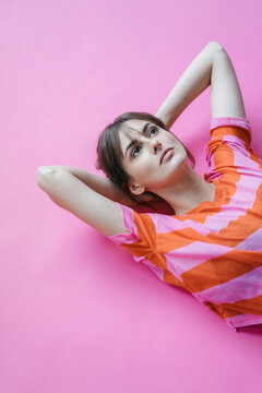 Woman lying on pink background with hands behind head, thinking