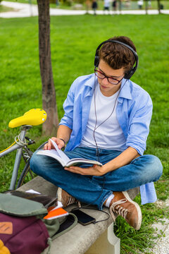 Young man with racing cycle and headphones sitting on a bench reading book