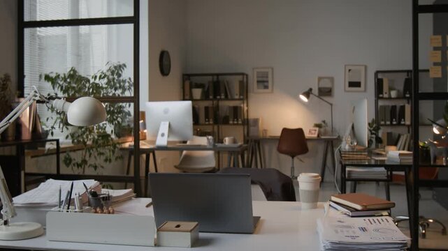 No people shot of modern office interior with several workstations, and white desk with laptop, documents, and lamp
