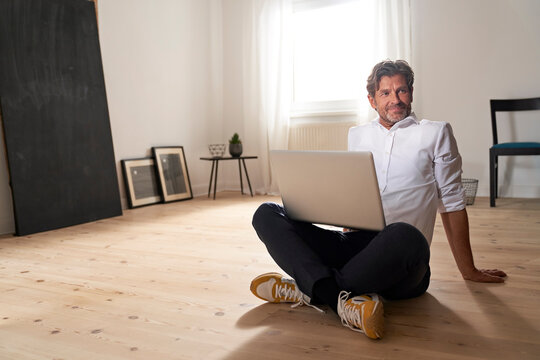 Portrait of smiling mature man with laptop sitting on the floor at home