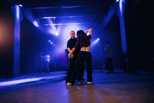 Two dancers performing in a dimly lit venue with colorful stage lights, showcasing dynamic movement and stylish outfits during a dance event or class