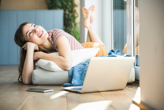 Relaxed young woman lying at the window at home with laptop