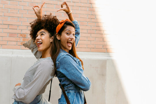 Cheerful friends pointing towards each other while standing back to back against wall