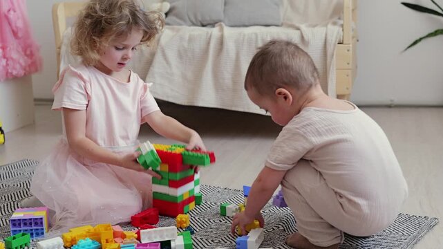 Two young children are engaged in play with colorful building blocks on the floor. One child builds a tower while the other focuses on stacking blocks